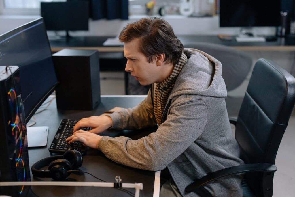 A man in a grey hoodie sites at a desk and types on a computer keyboard.